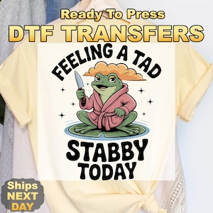 May include: A light yellow t-shirt with a graphic of a frog wearing a pink robe and holding a knife. The text reads "Feeling a tad stabby today". Above the graphic is the text "Ready To Press DTF TRANSFERS".