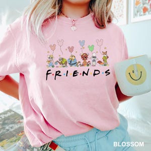 May include: Pink Friends graphic t-shirt featuring cartoon characters holding Mickey Mouse-shaped balloons. The word "FRIENDS" is printed in black with colorful dots. A white mug with a yellow smiley face is held in the image.