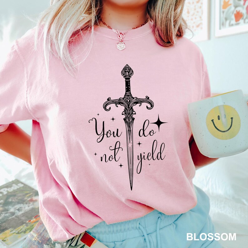 Peut inclure: T-shirt rose clair avec une &eacute;p&eacute;e noire et l'inscription "You do not yield". Le t-shirt est associ&eacute; &agrave; un short bleu clair et une tasse blanche avec un smiley jaune.
