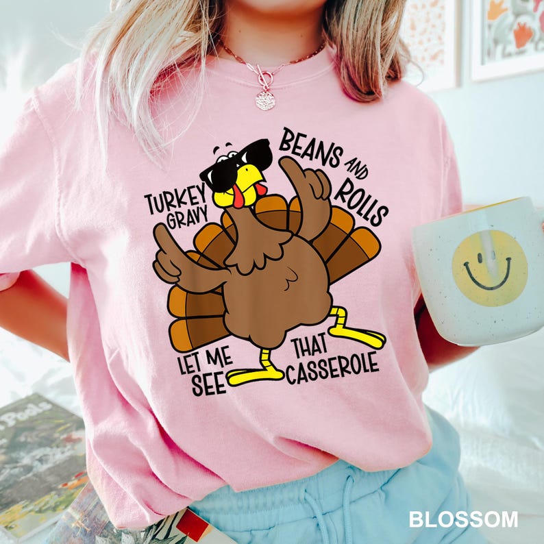 May include: Pink sweatshirt with a cartoon turkey wearing sunglasses and the text "Turkey Gravy, Beans and Rolls, Let Me See That Casserole." A white mug with a yellow smiley face is held in the hand.