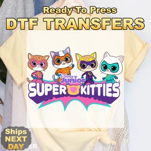 May include: A light yellow t-shirt featuring a graphic of four cartoon kittens in superhero costumes. The text "Ready To Press DTF TRANSFERS" is at the top, and "Disney Junior SUPER KITTIES" is at the bottom.