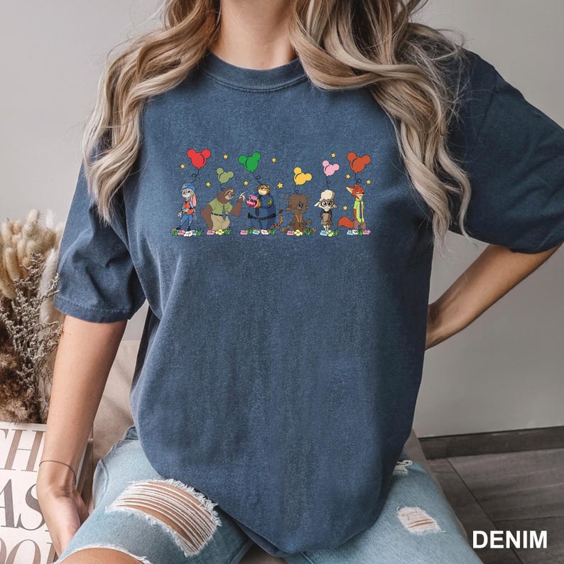 May include: Denim blue t-shirt with a graphic of cartoon characters holding heart-shaped balloons. The characters are in a row, with floral accents. The word "DENIM" is visible in the bottom right corner of the image.