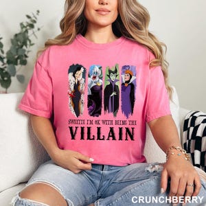 Puede incluir: Camiseta rosa con cuatro villanos de Disney: Cruella de Vil, &Uacute;rsula, Mal&eacute;fica y la Reina Malvada. El texto "Sweetie I'm ok with being the Villain" est&aacute; impreso debajo de los personajes en negro.