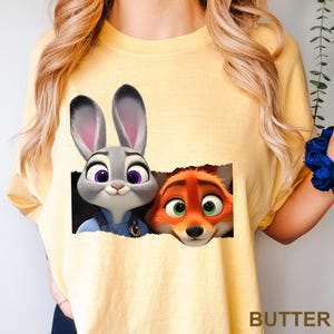 May include: Yellow t-shirt featuring a graphic of Judy Hopps and Nick Wilde from Zootopia. Judy, a gray rabbit, is in a police uniform. Nick, an orange fox, has green eyes. The word "BUTTER" is printed in the bottom right corner.