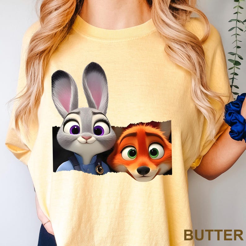 May include: Yellow t-shirt featuring a graphic of Judy Hopps and Nick Wilde from Zootopia. Judy, a gray rabbit, is in a police uniform. Nick, an orange fox, has green eyes. The word "BUTTER" is printed in the bottom right corner.
