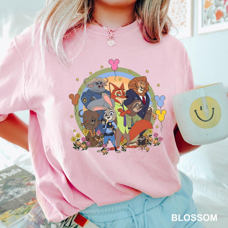 Pode incluir: Camiseta rosa claro com um gr&aacute;fico colorido de personagens de Zootopia, incluindo Judy Hopps e Nick Wilde, rodeados por flores e estrelas. A camiseta tem gola redonda e mangas curtas. Uma caneca branca com um rosto sorridente amarelo &eacute; segurada na m&atilde;o.