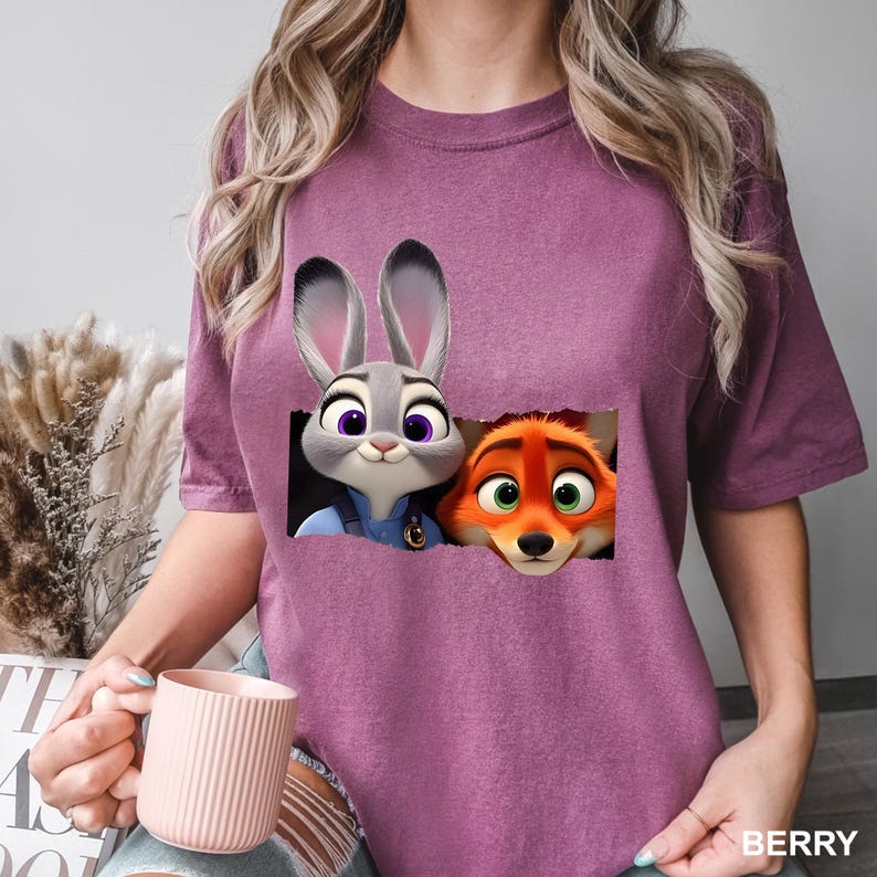 May include: Berry-colored t-shirt featuring a graphic of Judy Hopps and Nick Wilde from Zootopia. The cartoon characters are in a rectangular design. The shirt has short sleeves and a crew neck.