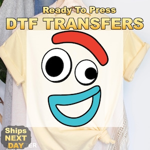 May include: A light yellow t-shirt featuring a cartoon face with a red eyebrow, two black and white eyes, and a blue mouth. The text "Ready To Press DTF TRANSFERS" is at the top. The shirt is knotted at the bottom.
