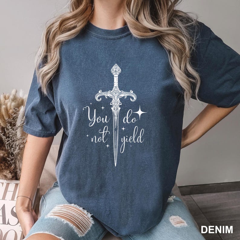 Peut inclure: T-shirt bleu denim avec un motif blanc d'&eacute;p&eacute;e et le texte "You do not yield". Le t-shirt a des manches courtes et une coupe ample. Le mot "DENIM" est imprim&eacute; en bas &agrave; droite.
