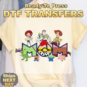 May include: A light yellow t-shirt featuring a DTF transfer design with the text "Ready To Press DTF TRANSFERS". The design includes cartoon characters and the word "MOM".
