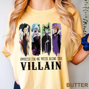 Puede incluir: Camiseta amarilla mantequilla con cuatro personajes de villanos de dibujos animados en marcos rectangulares verticales. El texto "Sweetie I'm ok with being the Villain" est&aacute; impreso debajo en negro.