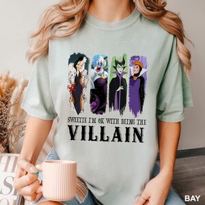 Puede incluir: Camiseta verde claro con una ilustraci&oacute;n de cuatro villanos de dibujos animados y el texto "Sweetie I'm OK with being the Villain". La camiseta tiene cuello redondo y mangas cortas.