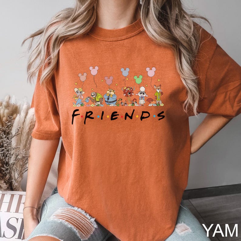 May include: Burnt orange t-shirt featuring the word "FRIENDS" in a colorful font, with cartoon characters and Mickey Mouse balloon designs above the text. The shirt is a comfortable, casual style.