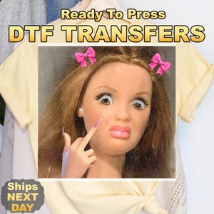 May include: A DTF transfer design featuring a doll-like face with exaggerated features, pink bows, and a raised finger. The text "Ready To Press DTF TRANSFERS" is displayed above the image. The shirt reads "Ships NEXT DAY".