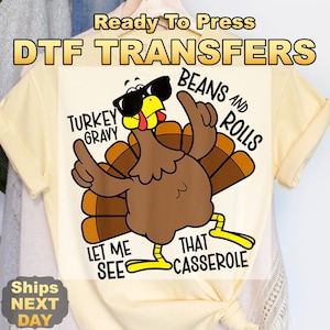 May include: A cream-colored DTF transfer featuring a cartoon turkey wearing sunglasses and gesturing with its wings. Text includes "Turkey Gravy", "Beans and Rolls", "Let Me See", and "That Casserole". The words "Ready To Press" and "DTF TRANSFERS" are at the top.