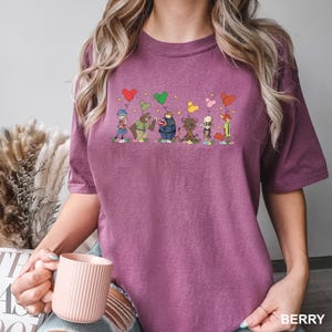 May include: Berry-coloured t-shirt with a cartoon design of animal characters holding heart-shaped balloons. The characters stand on flowers. The word "BERRY" is printed in white at the bottom right of the shirt.