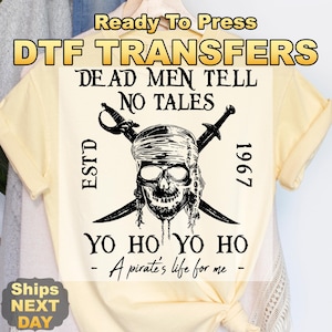 May include: A light yellow t-shirt featuring a pirate skull design with crossed swords. The design includes the text "DEAD MEN TELL NO TALES", "YO HO YO HO - A pirate's life for me -", and "ESTD 1967". The words "Ready To Press" and "DTF TRANSFERS" are at the top.