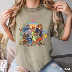 Pode incluir: Camiseta c&aacute;qui com um gr&aacute;fico colorido de personagens do filme de anima&ccedil;&atilde;o Zootopia. O design apresenta Judy Hopps, Nick Wilde e outros personagens, rodeados por flores e bal&otilde;es.