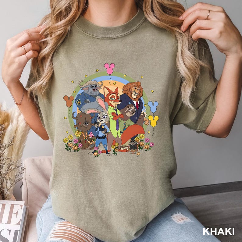 Pode incluir: Camiseta c&aacute;qui com um gr&aacute;fico colorido de personagens do filme de anima&ccedil;&atilde;o Zootopia. O design apresenta Judy Hopps, Nick Wilde e outros personagens, rodeados por flores e bal&otilde;es.