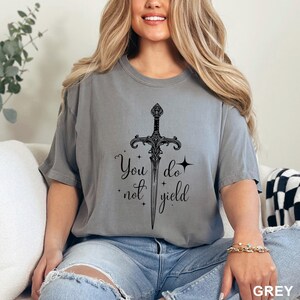 Peut inclure: T-shirt gris avec un motif d'&eacute;p&eacute;e noire et l'inscription "You do not yield". L'&eacute;p&eacute;e est d&eacute;taill&eacute;e et centr&eacute;e. Le t-shirt est de couleur grise unie.