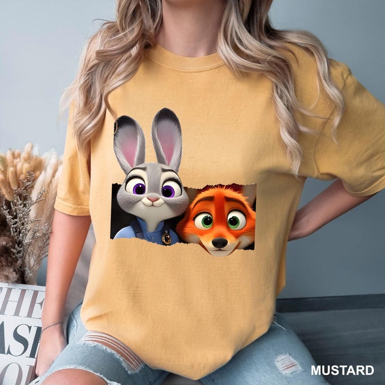 May include: Mustard-colored t-shirt featuring a graphic of Judy Hopps and Nick Wilde from Zootopia. Judy, a gray rabbit, and Nick, an orange fox, are depicted peeking out from a torn rectangle. The word "MUSTARD" is printed at the bottom.