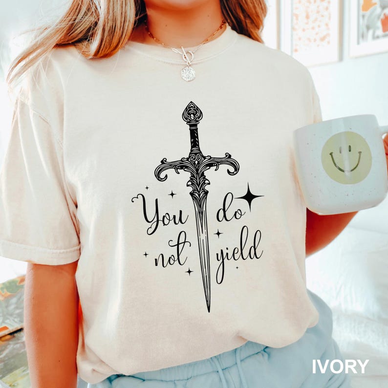 Peut inclure: T-shirt ivoire avec un dessin d'&eacute;p&eacute;e noire et le texte "You do not yield". Le motif comprend des &eacute;toiles et des &eacute;l&eacute;ments d&eacute;coratifs. Une tasse blanche avec un smiley est &eacute;galement visible.