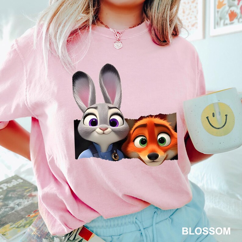 May include: A light pink t-shirt featuring a graphic of Judy Hopps and Nick Wilde from Zootopia. The characters are peeking out of a torn rectangle. A light blue mug with a yellow smiley face is held in the image.