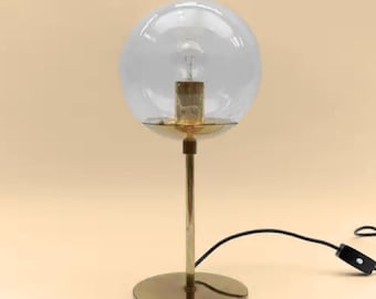 May include: A vintage-style table lamp with a clear, spherical glass shade. The lamp features a brass-colored base, stem, and a circular reflector. A black power cord extends from the base. The lamp is designed to hold a single light bulb.
