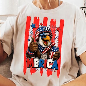 May include: A cream-coloured t-shirt featuring a graphic of a bald eagle wearing a helmet, star-shaped sunglasses, and a bandana. The eagle gives a thumbs up while holding a drink. The text reads USA and MERICA, set against red stripes.