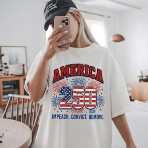 Puede incluir: Una camiseta blanca de manga corta con un gráfico que muestra el texto rojo AMERICA sobre el número 250, relleno con un patrón de estrellas y rayas. Fuegos artificiales rodean el número y el texto IMPEACH. CONVICT. REMOVE. está debajo.