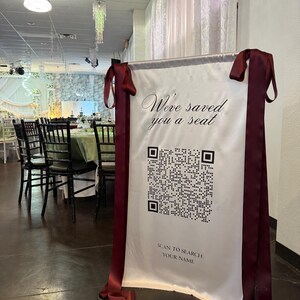 May include: A white sign with the text "We've saved you a seat" and a QR code. The sign is framed with a dark red ribbon and is displayed on a black metal stand. The background shows a banquet hall with tables and chairs.