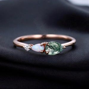 Könnte beinhalten: Roségoldener Ring mit drei Edelsteinen: einem kleinen runden Diamanten, einem birnenförmigen Opal und einem größeren birnenförmigen Moosachat. Der Ring ist vor einem dunklen Hintergrund platziert, der die Farben und Details hervorhebt.