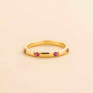 Könnte beinhalten: Ein goldfarbener Ring mit einer Reihe kleiner, rechteckiger Goldsegmente, die jeweils von einem leuchtenden, runden, roten Edelstein durchbrochen sind. Der Ring ist vor einem weichen, neutralen Hintergrund platziert, der sein elegantes Design hervorhebt.