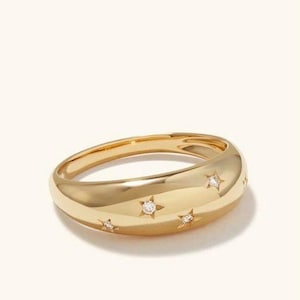 Könnte beinhalten: Ein goldfarbener Ring mit einem abgerundeten Band und einer Reihe kleiner sternförmiger Akzente, die jeweils einen kleinen, klaren Stein enthalten. Der Ring hat eine glatte, polierte Oberfläche. Schmuck.
