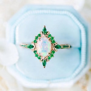 May include: An opal and emerald ring set in gold. The oval opal is surrounded by a halo of small green emeralds, with emerald accents on the band. The ring is displayed in a light blue ring box.