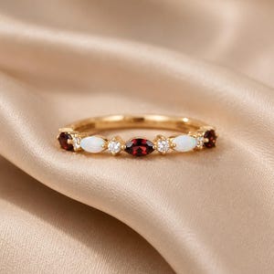 Könnte beinhalten: Ein Goldring mit einem abwechselnden Muster aus markisengeschliffenen dunkelroten Granaten, ovalen weißen Opalen und kleinen runden klaren Steinen. Die Steine sind in einer Reihe entlang der oberen Hälfte des dünnen, polierten Goldrings gefasst.