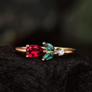 Könnte beinhalten: Ein goldener Ring mit einem birnenförmigen, tiefroten Edelstein und zwei grünen, blattförmigen Steinen. Ein kleiner, klarer, runder Stein ist in der Nähe der Blätter gesetzt. Der Ring liegt auf einer dunklen, strukturierten Oberfläche.