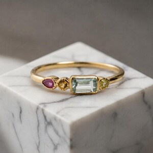 Könnte beinhalten: Ein Goldring mit einem zentralen, rechteckigen hellblauen Edelstein, flankiert von einem kleinen runden gelben Stein, einem birnenförmigen rosa Stein und einem kleinen dreieckigen grünen Stein. Der Ring ist schmal und poliert mit mehreren Steinen.