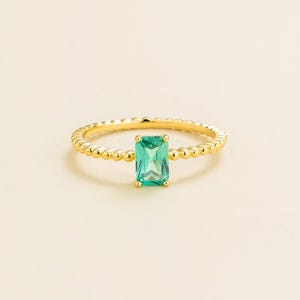 Könnte beinhalten: Ein goldener Ring mit einem rechteckigen, smaragdgeschliffenen, hellgrünen Edelstein. Das Band hat ein Perlendesign. Der Ring ist vor einem neutralen Hintergrund platziert, der die Details des Schmucks hervorhebt.