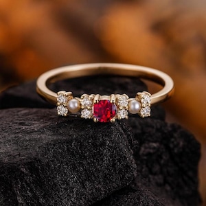 Könnte beinhalten: Ein goldener Ring mit einem quadratisch geschliffenen roten Edelstein in der Mitte, flankiert von kleinen Perlen und Diamantakzenten. Der Ring hat ein dünnes, poliertes Goldband. Der Ring wird auf einer dunklen, strukturierten Oberfläche präsentiert.