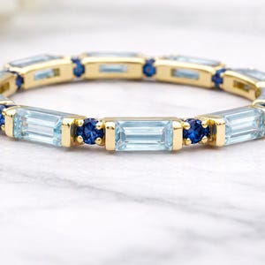 Könnte beinhalten: Ein goldener Ring mit abwechselnd hellblauen und dunkelblauen Edelsteinen. Die hellblauen Steine sind rechteckig, während die dunkelblauen Steine rund sind. Der Ring liegt auf einer weißen Marmoroberfläche.