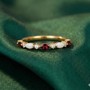 May include: A gold ring with alternating gemstones, including oval-cut red garnets, white opals, and small round diamonds. The ring is set against a dark green background.