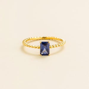 May include: A gold ring with a rectangular, dark blue gemstone. The band features a beaded design. The ring is set against a cream-colored background, highlighting the jewelry's details. A simple and elegant piece.