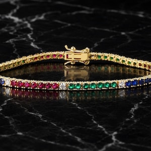 Könnte beinhalten: Ein goldenes Tennisarmband mit einer durchgehenden Reihe rund geschliffener Edelsteine in einer lebendigen Regenbogenfarbpalette, darunter rote, grüne, blaue und klare Steine. Das Armband wird mit einem goldenen Kastenschloss verschlossen.
