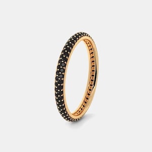Könnte beinhalten: Ein goldfarbener Ring mit einem Band aus kleinen, schwarzen Edelsteinen. Der Ring ist vor einem schlichten, weißen Hintergrund platziert, der sein Design hervorhebt. Die Edelsteine sind gleichmäßig um den Ring herum angeordnet und bilden ein durchgehendes Muster.