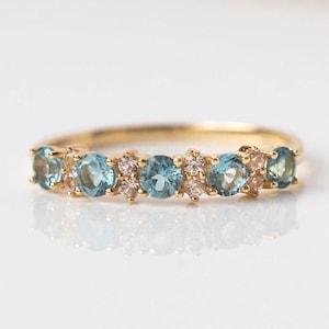 Könnte beinhalten: Ein goldener Ring mit einer Reihe von hellblauen, runden Edelsteinen, die mit kleineren, klaren und rosa Edelsteinen abwechseln. Der Ring ist vor einem weißen Hintergrund platziert, um die Details hervorzuheben.