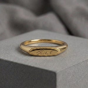 Puede incluir: Un anillo de tono dorado que presenta una cara de forma ovalada con un borde de cuentas. El centro del óvalo está grabado con la palabra "Bitch" en una fuente de escritura cursiva. La banda es lisa y pulida, manteniendo un ancho constante.
