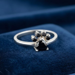 Könnte beinhalten: Ein silberner Ring mit einem Pfotenabdruck-Design. Der Ring hat ein Band und einen Pfotenabdruck aus schwarzen Edelsteinen. Der Pfotenabdruck hat einen großen, dreieckigen Mittelstein und vier kleinere Steine darüber.