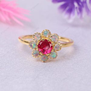 Könnte beinhalten: Ein goldener Ring mit einem blütenförmigen Design. Der Mittelstein ist ein leuchtendes Rot, umgeben von kleineren opalisierenden Steinen. Der Ring hat ein schlichtes, poliertes Goldband.