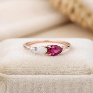 Könnte beinhalten: Ein roségoldener Ring mit drei Edelsteinen. Ein kleiner, runder, klarer Stein folgt einem birnenförmigen weißen Opal und einem größeren, birnenförmigen, tiefroten Rubin. Der Ring liegt auf einer neutralfarbenen Oberfläche.